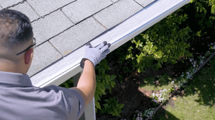 Person wearing gloves installs a mesh gutter guard along the edge of a shingled roof above a garden.