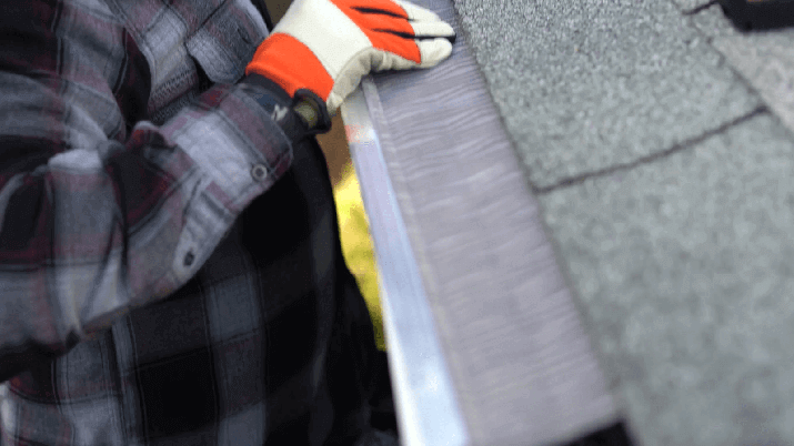Person wearing gloves installs metal flashing along the edge of a roof with asphalt shingles.