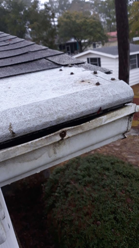 Close-up of a house roof edge and gutter with visible dirt, rust, and debris buildup on the surfaces.