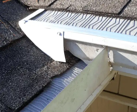 Close-up of a house roof corner showing a gutter guard installed over the rain gutter next to asphalt shingles.
