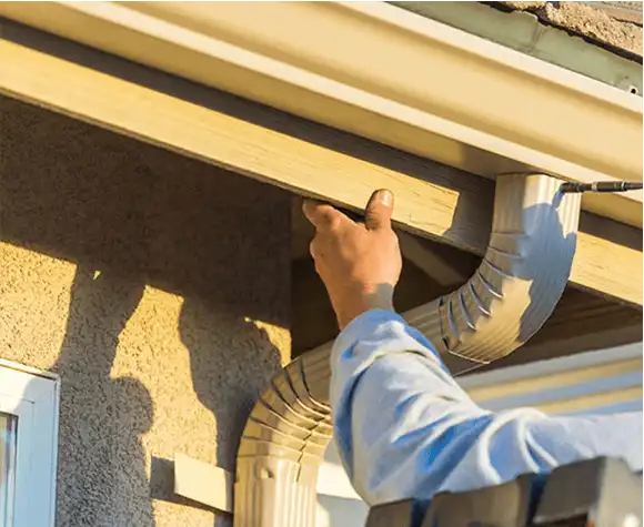 Person inspecting or repairing a house gutter and downspout attached to the roof edge on a sunny day.