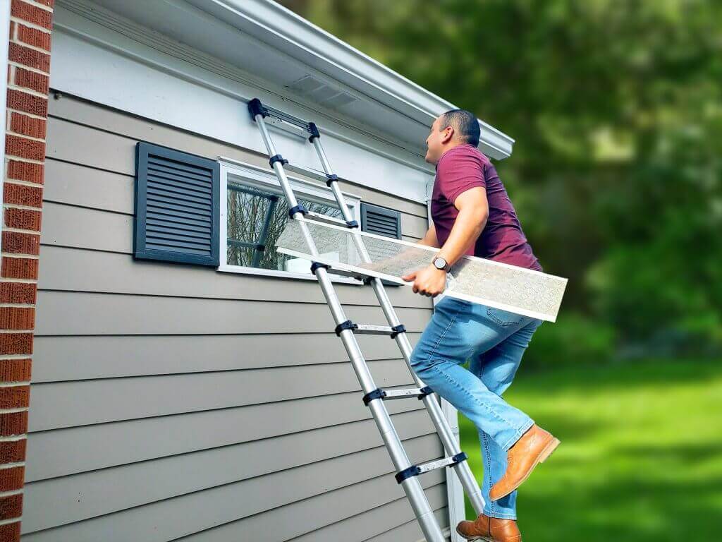 A man climbs a ladder to the roof of a house while holding a rectangular panel in his right hand.