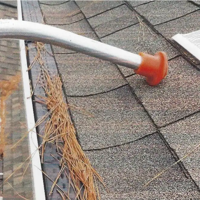 A leaf blower attachment is being used to clear pine needles from a house roof and rain gutter.