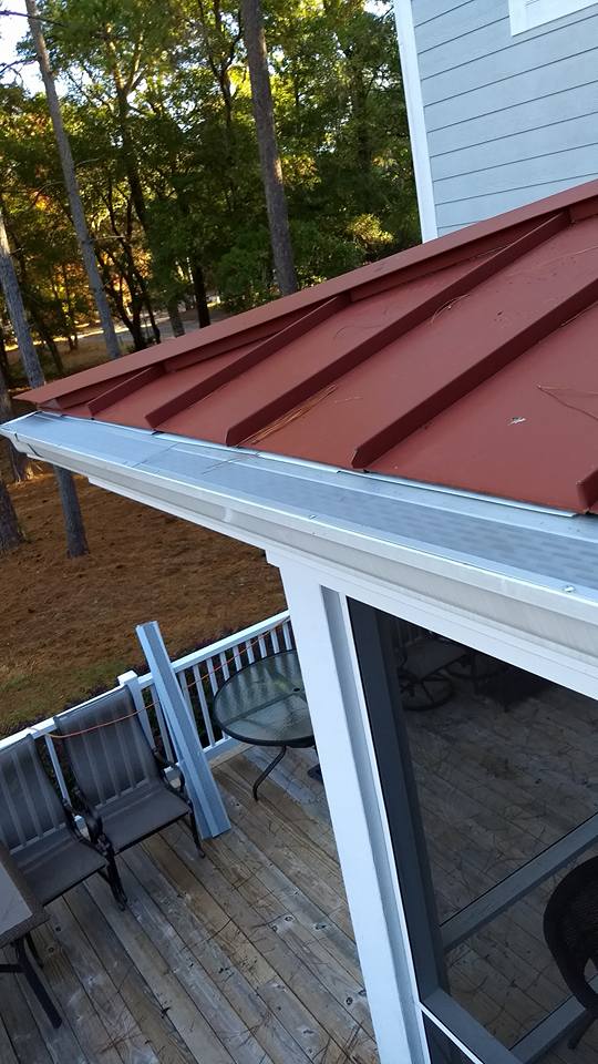 A red metal roof with a clean gutter above a wooden deck furnished with chairs and a glass table, surrounded by trees.