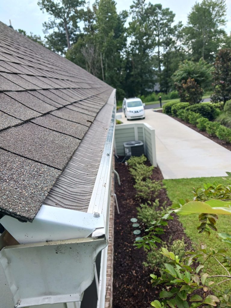 View of a house roof with gutter guards, a driveway, parked white van, and landscaped yard with trees and shrubs.