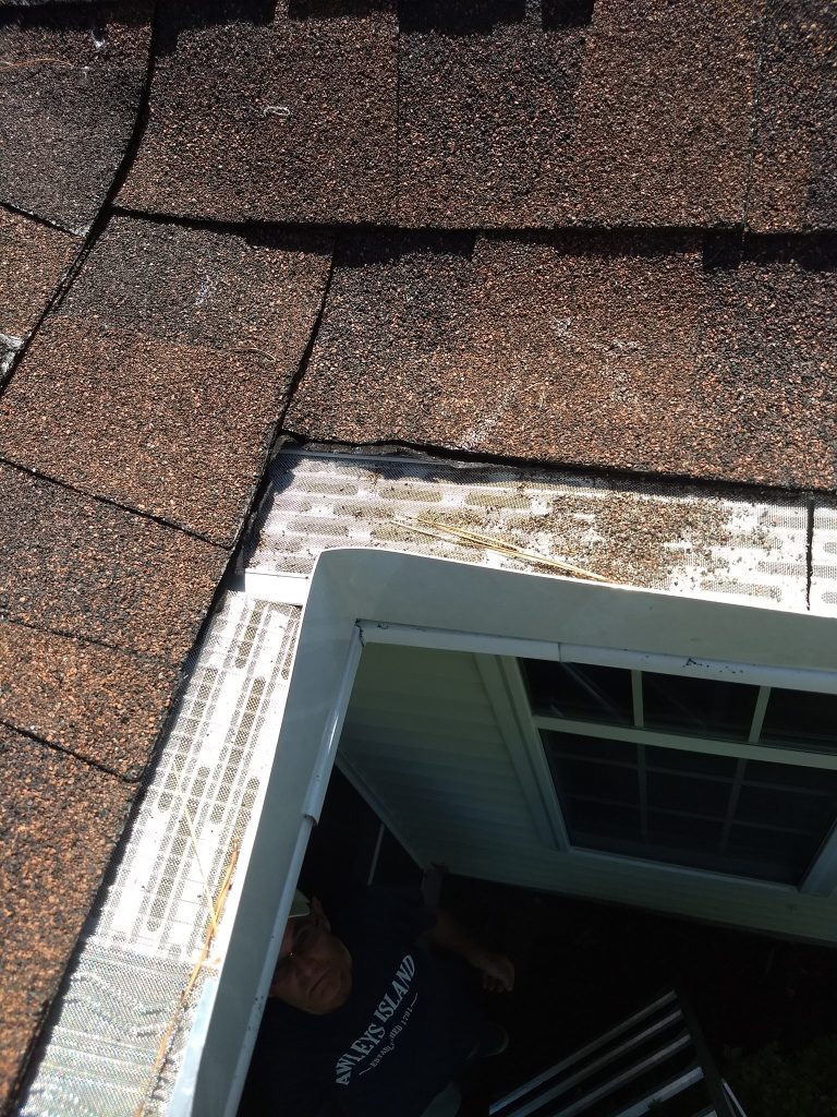 Close-up of a house roof corner with brown shingles, white gutter guards, and a person standing below.