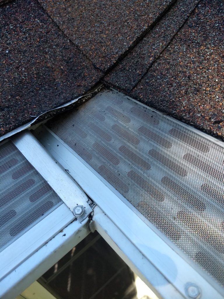 Close-up of a roof edge with shingles, metal mesh gutter guard, and part of a window frame visible.