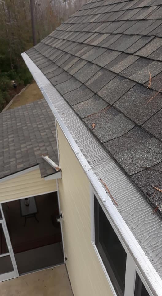 A house roof with gray shingles and a metal gutter guard, viewed from above, with some pine needles visible.