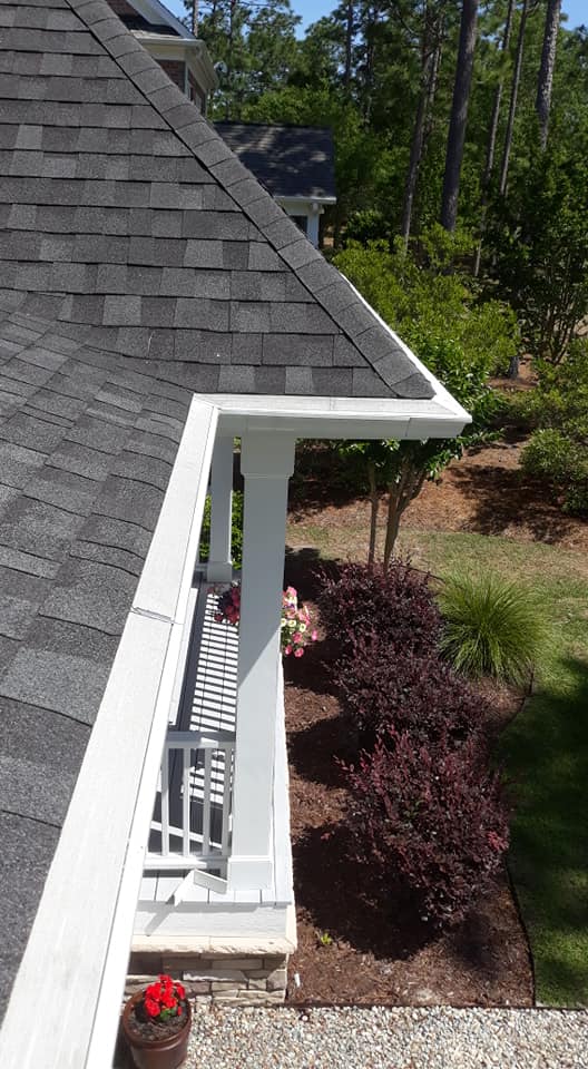 View of a house roof with clean gutters, white railing, shrubs, flowers, and a gravel pathway below, surrounded by greenery.