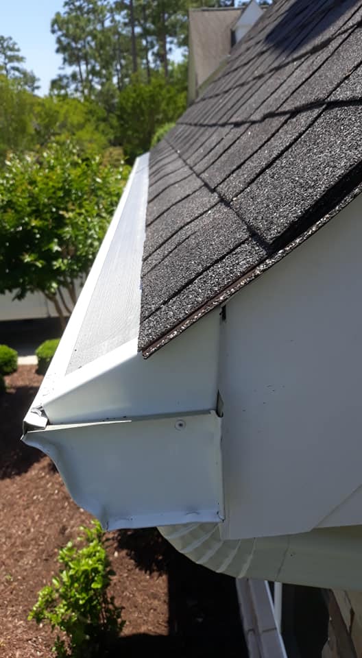 Close-up of a house roof edge with black shingles and white rain gutter in daylight.