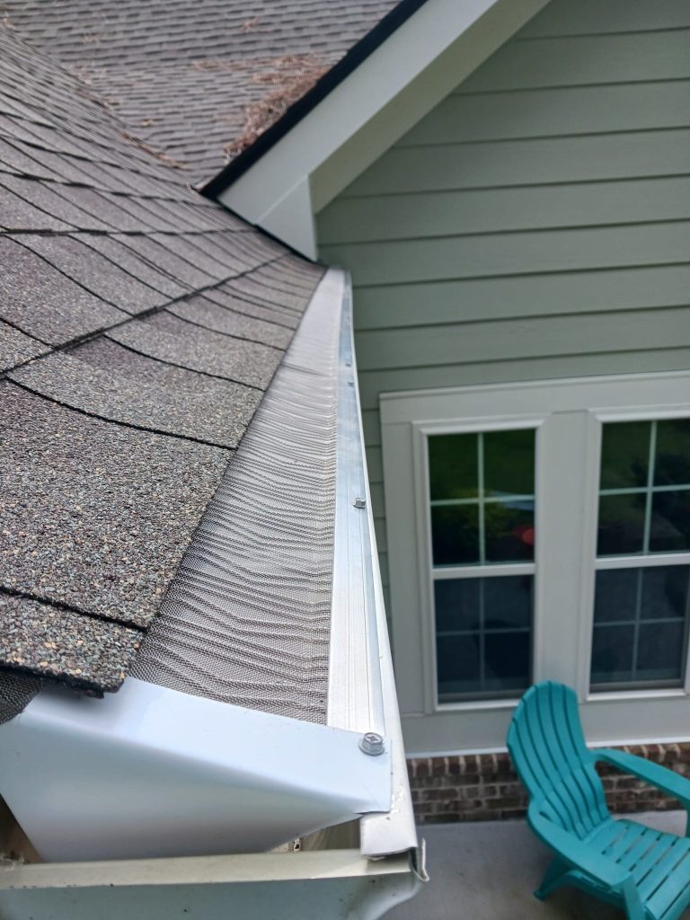 Close-up of a house roof showing a gutter with a metal mesh guard and part of a patio with blue chairs below.
